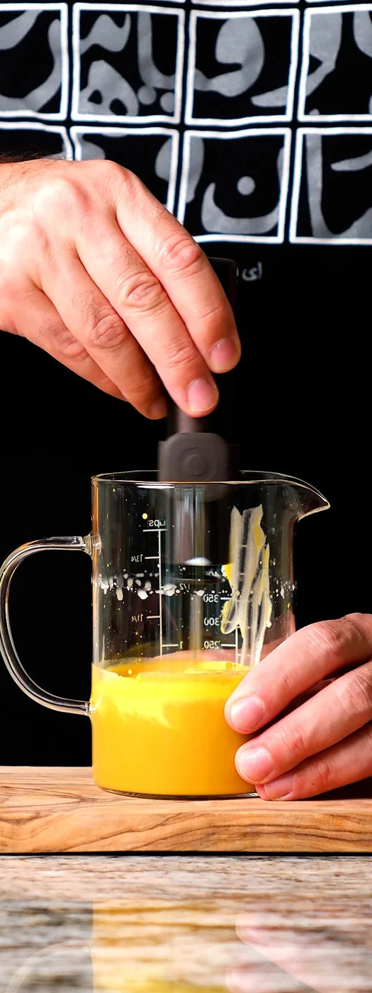 A handheld milk frother blending the golden saffron mixture in a glass jug